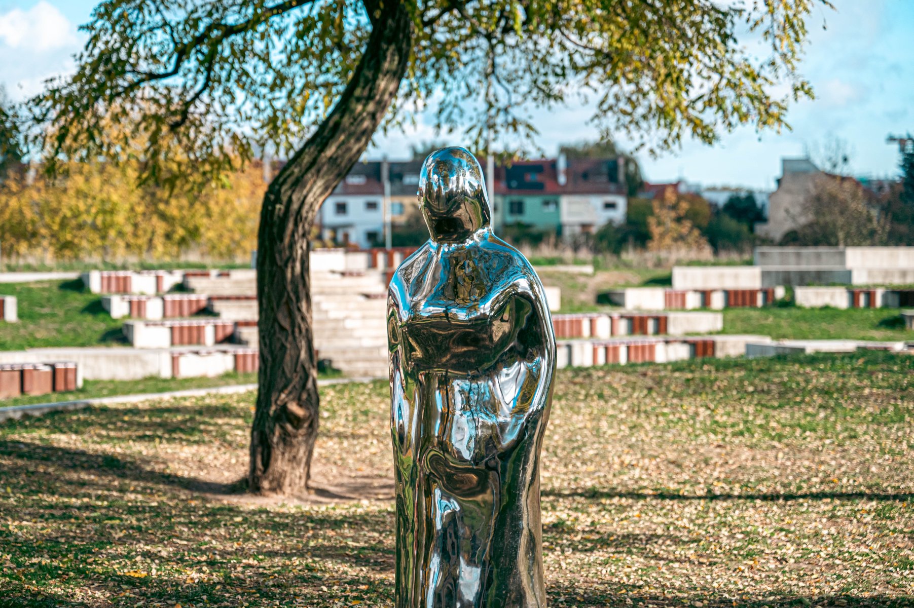 Metaliczna rzeźba postaci ludzkiej o refleksyjnej powierzchni, ustawiona w parku. Lustro odbija otoczenie, w tym drzewa z zielono-żółtymi liśćmi, co sugeruje jesienną porę roku. W tle widoczne są schodkowe tarasy oraz budynki, wskazujące na miejski parkowy krajobraz.