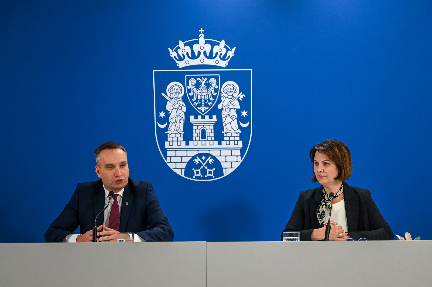 Mężczyzna i kobieta w strojach formalnych siedzą przy stole konferencyjnym, każdy przed mikrofonem. Mężczyzna przemawia. W tle, na niebieskiej ścianie, duży, biały herb miasta Poznania.