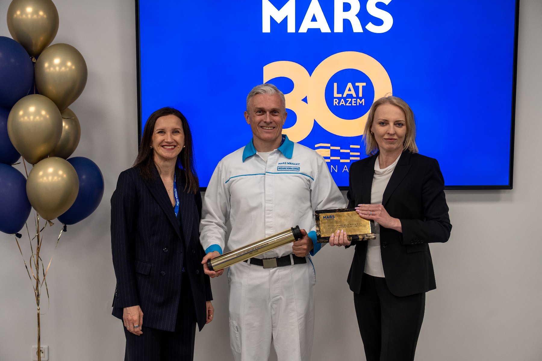 Mężczyzna w białym roboczym kombinezonie z niebieskimi akcentami oraz logo "Mars Wrigley" stoi w centrum kadru, trzymając podłużną, złotą tubę. Po jego lewej stronie stoi kobieta w ciemnym garniturze w prążki, a po prawej kobieta w czarnym żakiecie, która współtworzy uścisk na pamiątkowej, ciemnej statuetce ze złotą tabliczką.