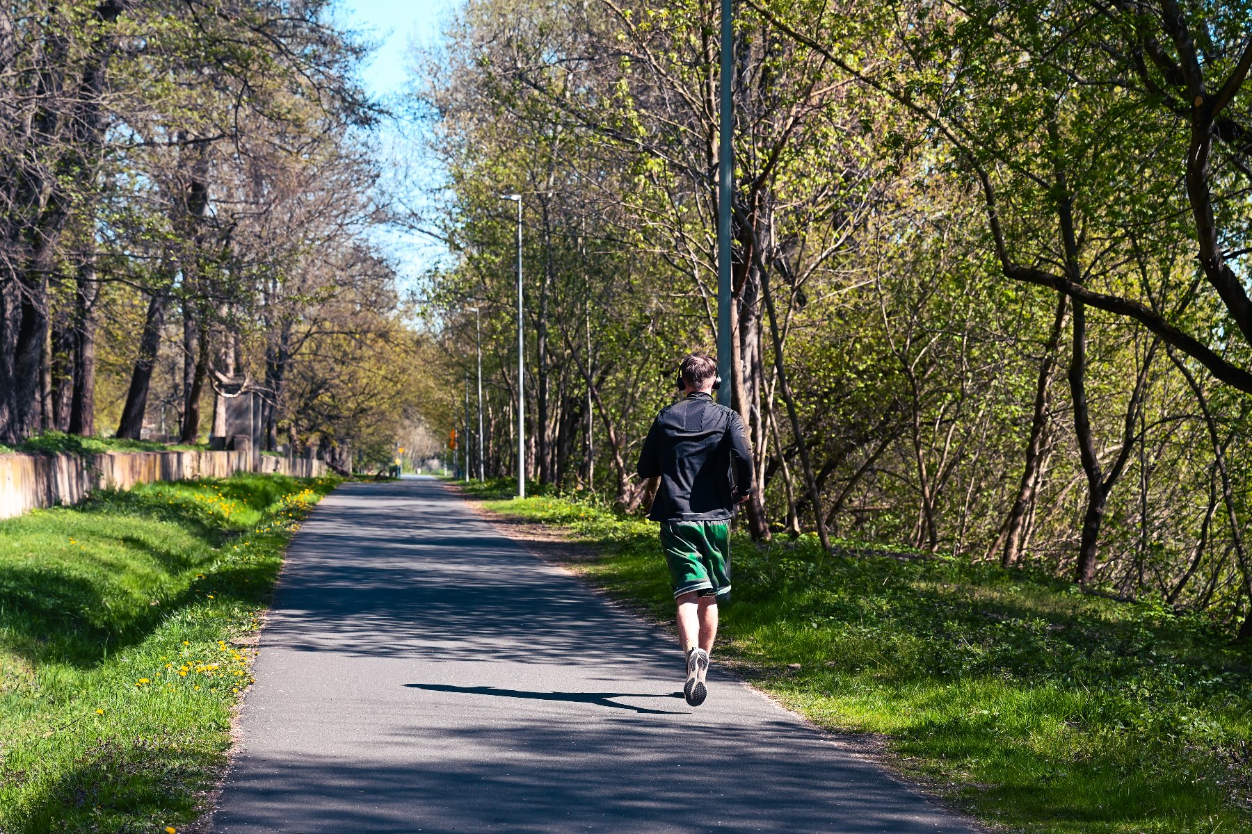 Mężczyzna w czarnej bluzie, zielonych spodenkach i słuchawkach biegnie tyłem do obiektywu środkiem asfaltowej ścieżki. Droga ciągnie się przez park, otoczona trawą oraz gęstymi, zielonymi drzewami po obu stronach. Wzdłuż trasy widoczne są latarnie, a po lewej stronie w głębi niski, kamienny murek.