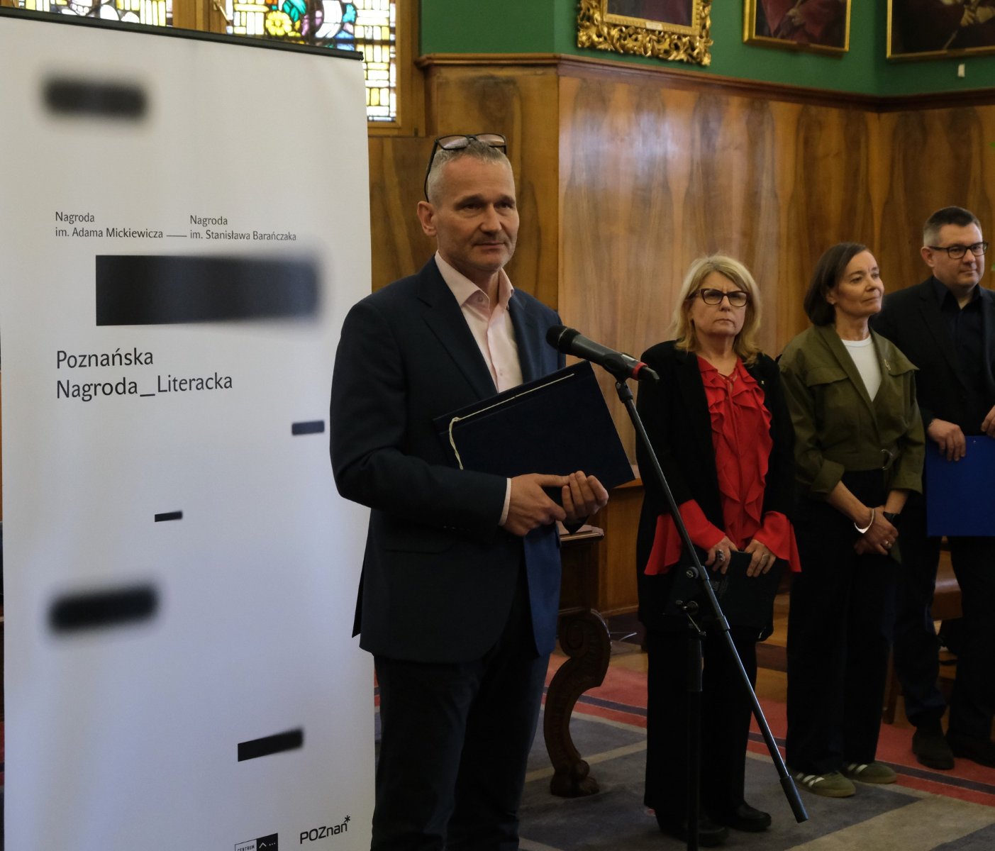 Mężczyzna w garniturze stoi przy mikrofonie i trzyma teczkę, przemawiając podczas oficjalnej uroczystości. Obok niego stoi kilka osób, a w tle widać drewniane ściany, obrazy oraz baner z napisem "Poznańska Nagroda Literacka"