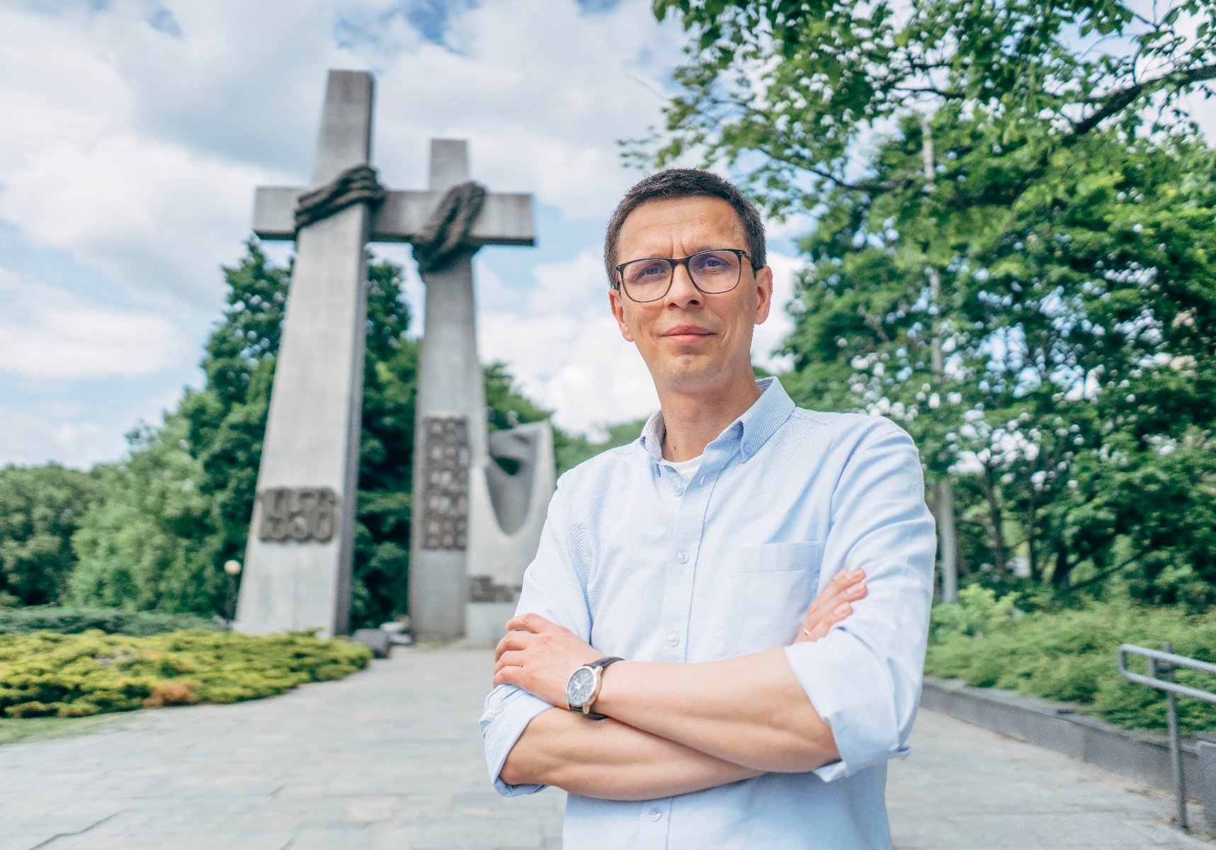 Mężczyzna w okularach i błękitnej koszuli pozuje na tle pomnika Poznańskich Krzyży na placu im. Adama Mickiewicza.