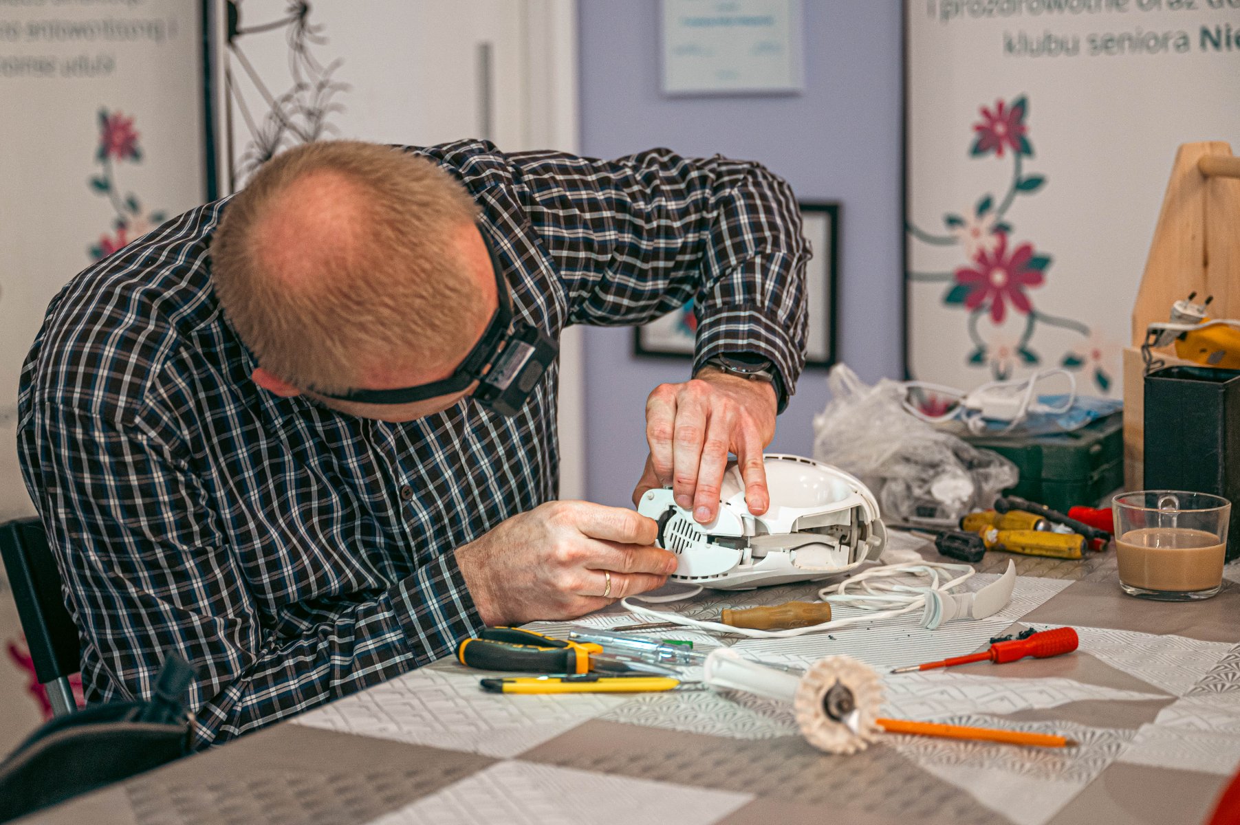 Mężczyzna w okularach na głowie pochyla się nad białym urządzeniem AGD, prawdopodobnie mikserem, które jest częściowo rozłożone. Na stole leżą liczne narzędzia. W tle widać fragmenty ścian i tablic.