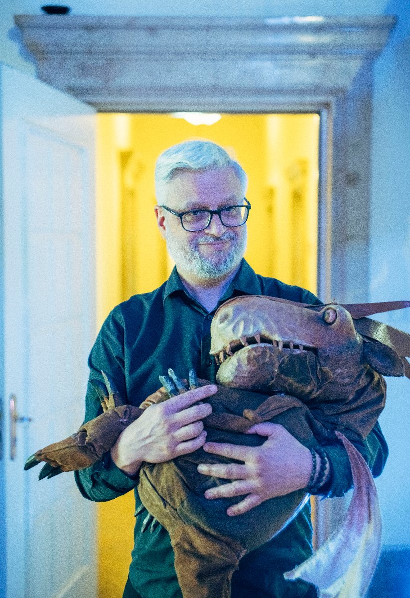 Mężczyzna z siwą brodą i w okularach trzyma w rękach teatralną lalkę-smoka.