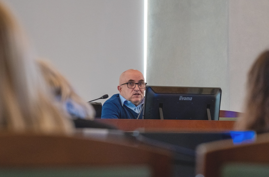 Mężczyzna za stołem prezydialnym przemawia w sali zwrócony do innych uczestników spotkania - więcej w treści artykułu - grafika artykułu