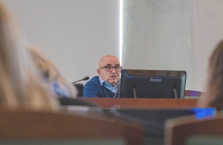 Mężczyzna za stołem prezydialnym przemawia w sali zwrócony do innych uczestników spotkania - więcej w treści artykułu
