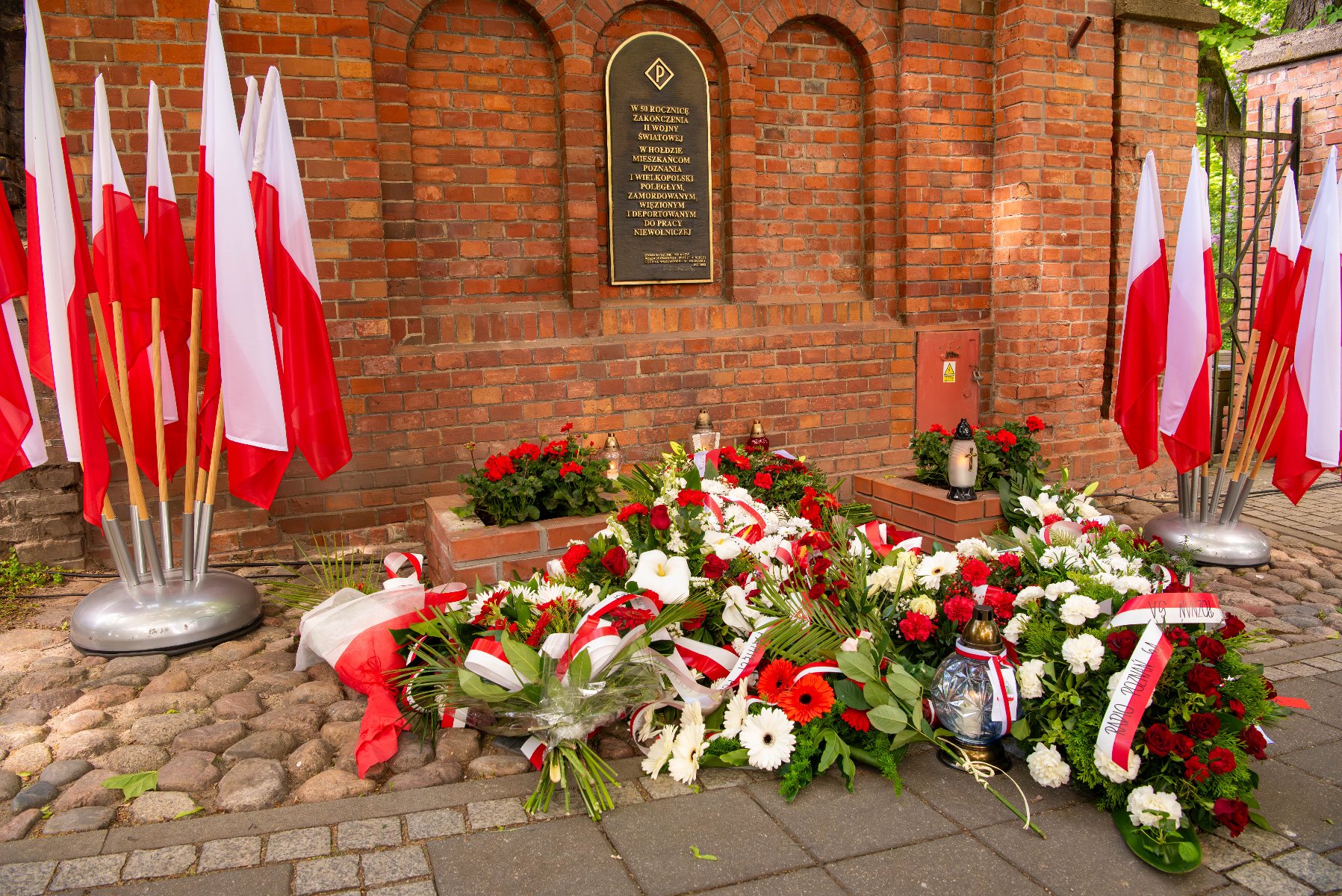 Miejsce pamięci z polskimi flagami oraz licznymi wiązankami kwiatów ułożonymi przed ceglaną ścianą. Na ścianie znajduje się tablica pamiątkowa z tekstem w języku polskim.