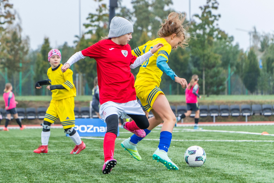 Młode piłkarki podczas meczu na zielonej murawie. Na pierwszym planie dziewczynka w czerwonej koszulce i szarej czapce walczy o piłkę z zawodniczką w żółto-niebieskim stroju. W tle widoczne inne piłkarki w żółtych i różowych koszulkach oraz niebieska tablica z białym napisem "POZnan". Za boiskiem rząd pustych krzeseł i drzewa.