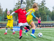 Młode piłkarki podczas meczu na zielonej murawie. Na pierwszym planie dziewczynka w czerwonej koszulce i szarej czapce walczy o piłkę z zawodniczką w żółto-niebieskim stroju. W tle widoczne inne piłkarki w żółtych i różowych koszulkach oraz niebieska tablica z białym napisem "POZnan". Za boiskiem rząd pustych krzeseł i drzewa.