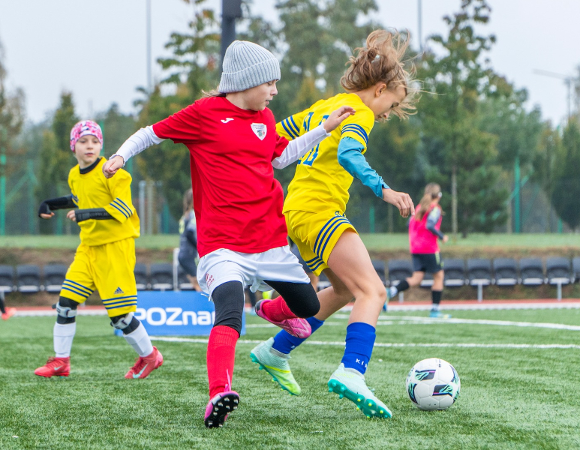 Młode piłkarki podczas meczu na zielonej murawie. Na pierwszym planie dziewczynka w czerwonej koszulce i szarej czapce walczy o piłkę z zawodniczką w żółto-niebieskim stroju. W tle widoczne inne piłkarki w żółtych i różowych koszulkach oraz niebieska tablica z białym napisem "POZnan". Za boiskiem rząd pustych krzeseł i drzewa.