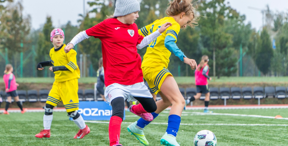 Młode piłkarki podczas meczu na zielonej murawie. Na pierwszym planie dziewczynka w czerwonej koszulce i szarej czapce walczy o piłkę z zawodniczką w żółto-niebieskim stroju. W tle widoczne inne piłkarki w żółtych i różowych koszulkach oraz niebieska tablica z białym napisem "POZnan". Za boiskiem rząd pustych krzeseł i drzewa.
