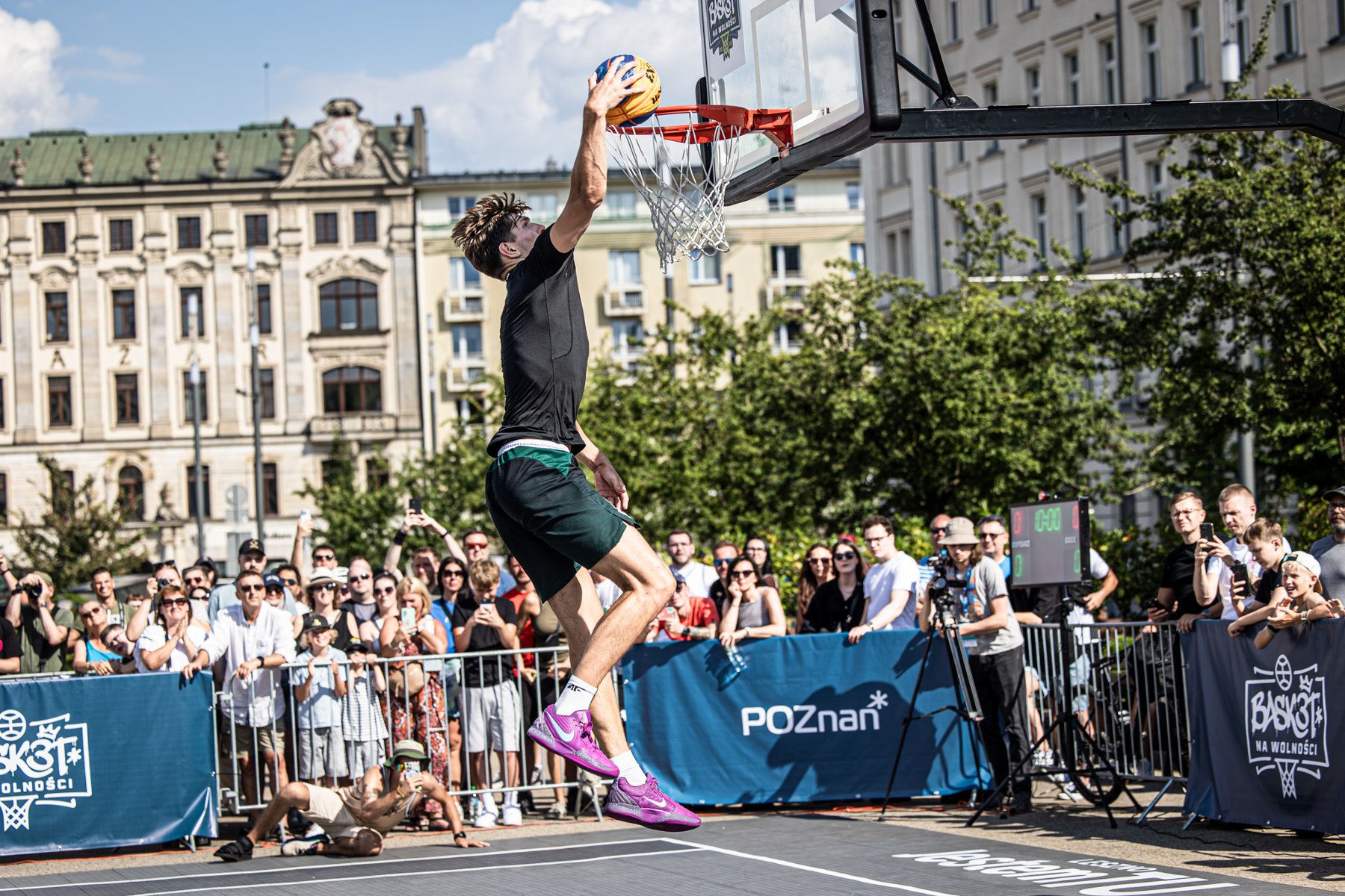 Młody koszykarz w czarnym stroju i fioletowych butach wykonuje wsad do kosza podczas turnieju koszykówki 3x3 "Basket na Wolności". Wydarzenie odbywa się na plenerowym boisku rozstawionym na placu Wolności w Poznaniu, w tle widać zgromadzoną publiczność, barierki z logo Poznania oraz zabytkową kamienicę Bazaru Poznańskiego.