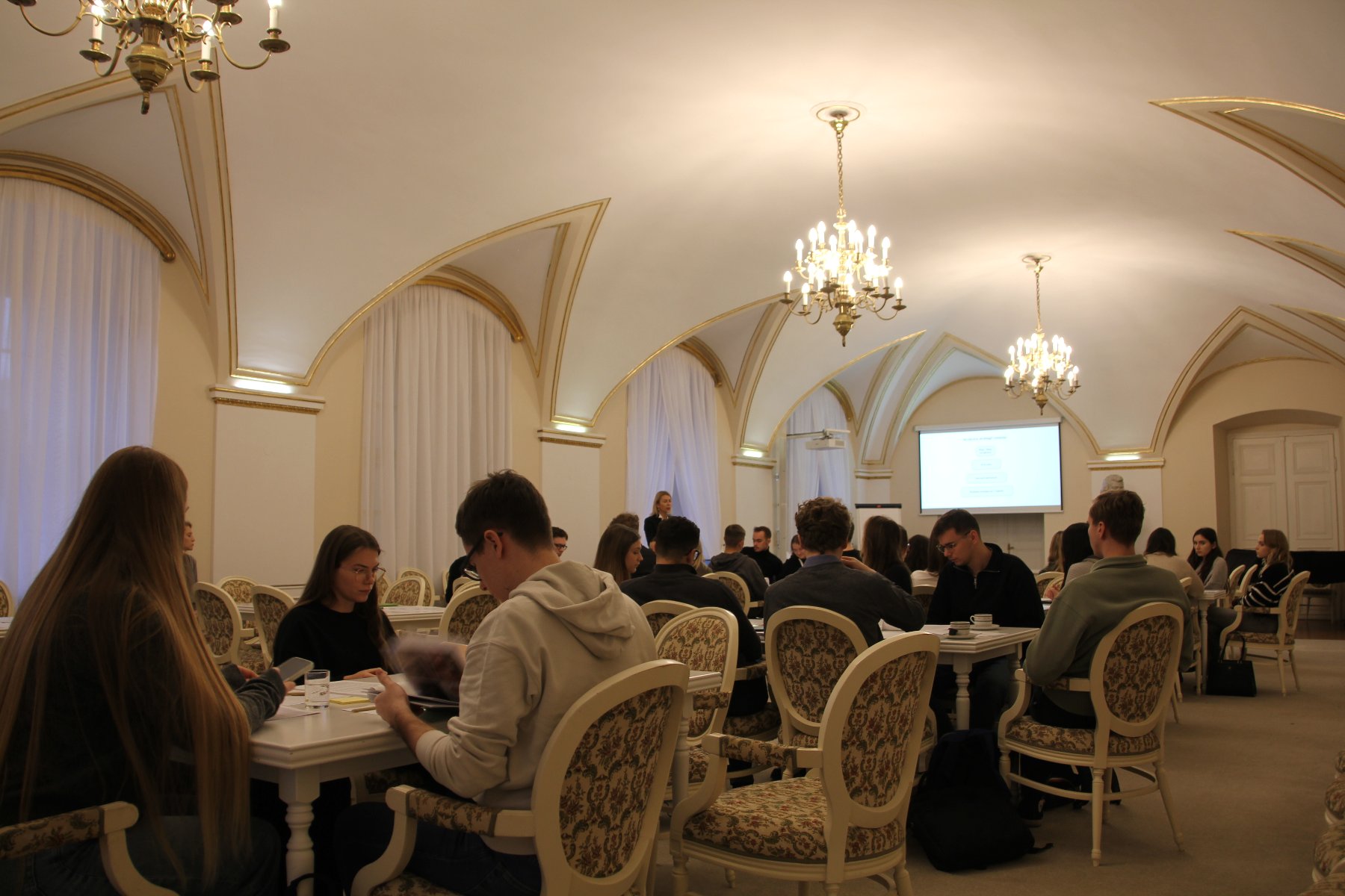 Młodzież przy kilku stolikach na sali urzędu, widok na tablicę do prezentacji