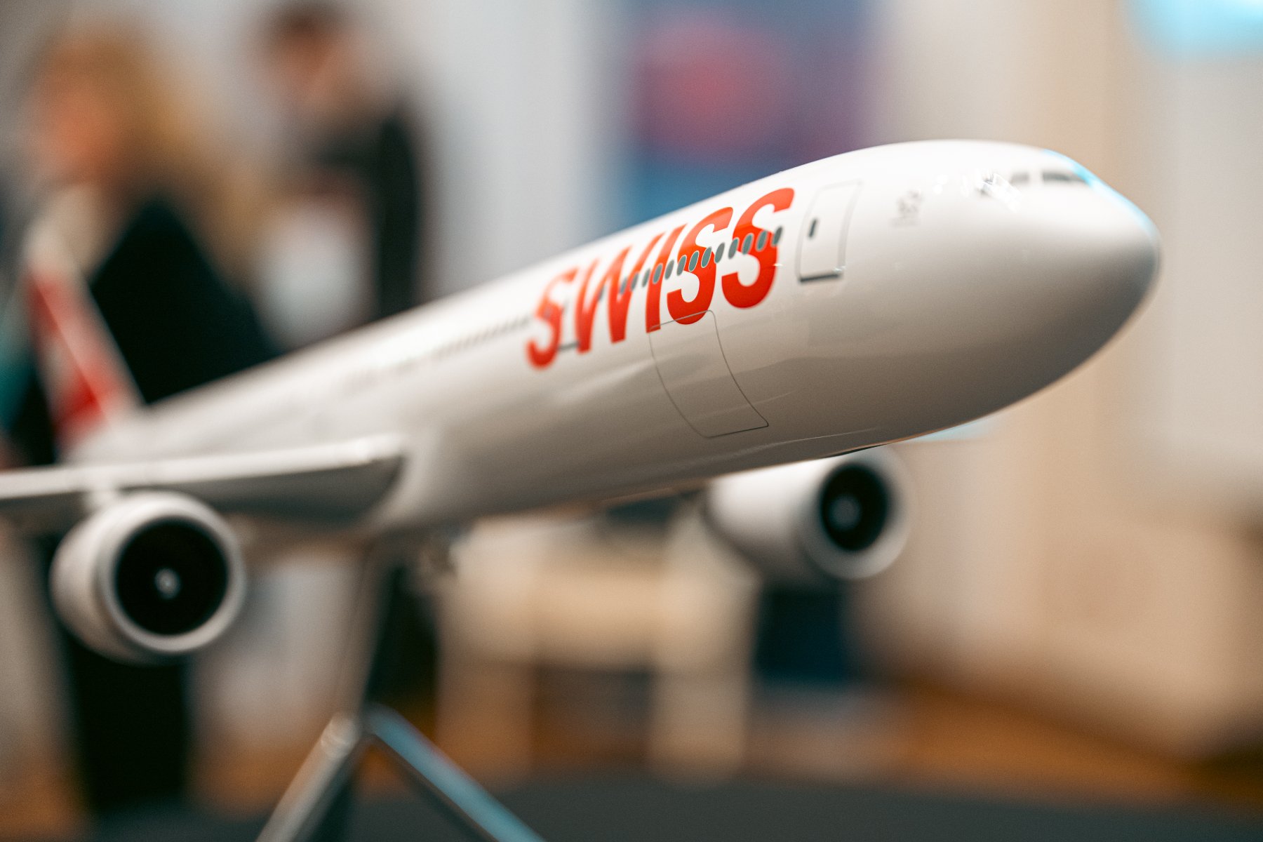 Model białego samolotu pasażerskiego na metalowym stojaku. Na kadłubie widoczny duży, czerwony napis "SWISS". Tło jest rozmyte, widać w nim zarysy postaci i wnętrza.