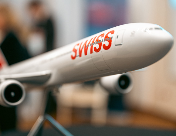 Model białego samolotu pasażerskiego na metalowym stojaku. Na kadłubie widoczny duży, czerwony napis "SWISS". Tło jest rozmyte, widać w nim zarysy postaci i wnętrza.