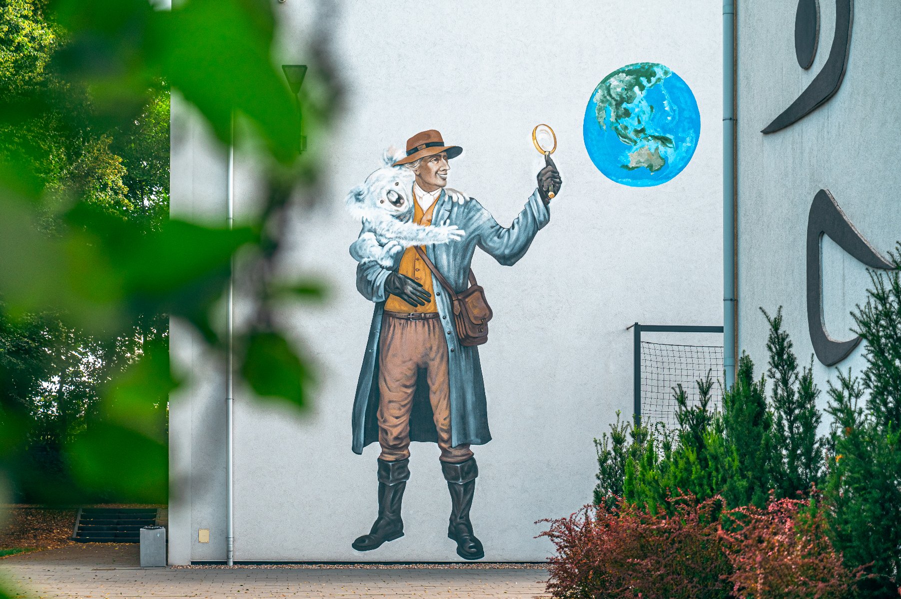 Mural na bocznej ścianie nowoczesnego budynku. Przedstawia on mężczyznę w stroju podróżnika (długi płaszcz, kapelusz, torba przewieszona przez ramię), który w jednej ręce trzyma białą koalę, a w drugiej unosi szkło powiększające, patrząc przez nie w stronę namalowanej obok kuli ziemskiej. Kula ziemska pokazuje kontynent australijski.