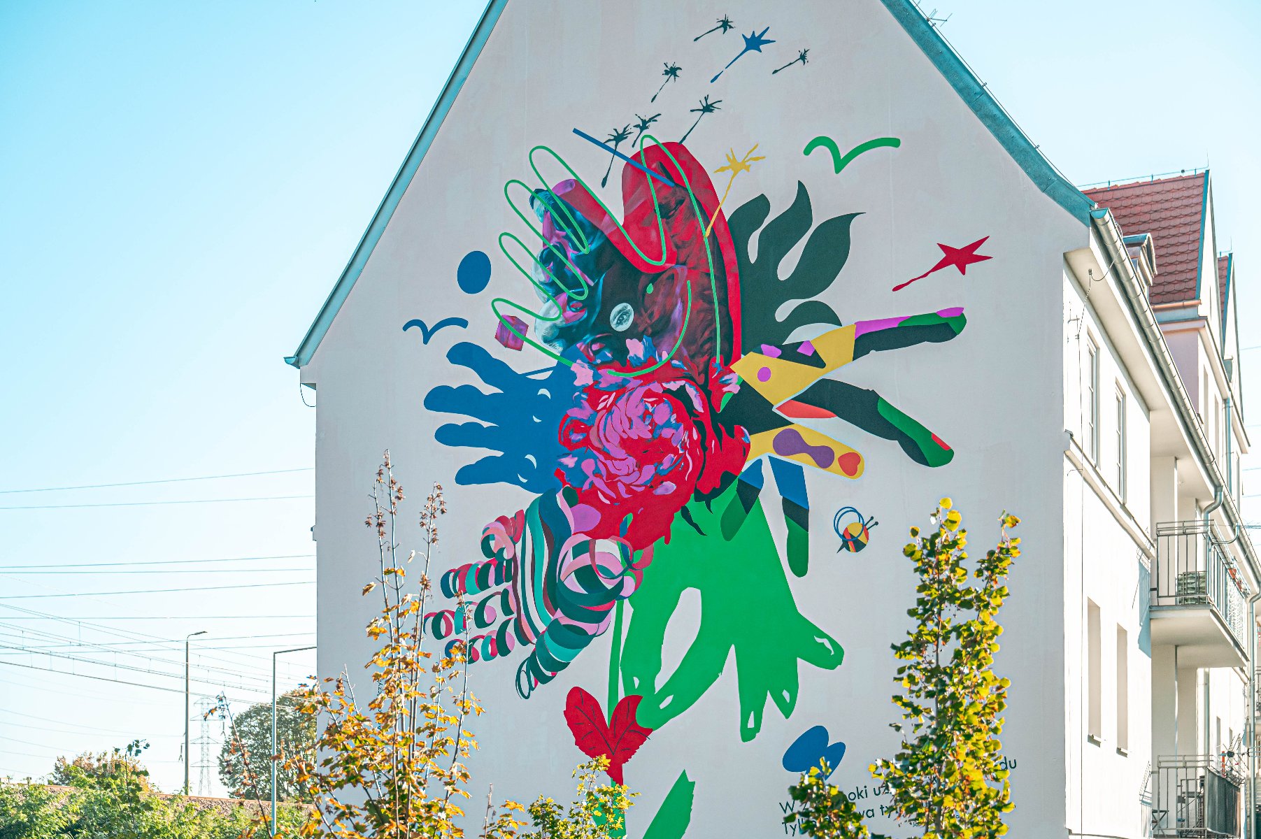 mural na zdjęciu kwiat, którego płatki tworzą dłonie symbolizujące cztery różne postawy artystyczne. Dłonie te nie tylko reprezentują wielogłosowość perspektyw twórców, lecz również różnorodność osób zaangażowanych w troskę o zieleń miejską, ogrody i parki Poznania. To symboliczne ujęcie jednoczy wszystkich tych, którzy działają na rzecz ekologii, niezależnie od wieku, profesji czy pochodzenia