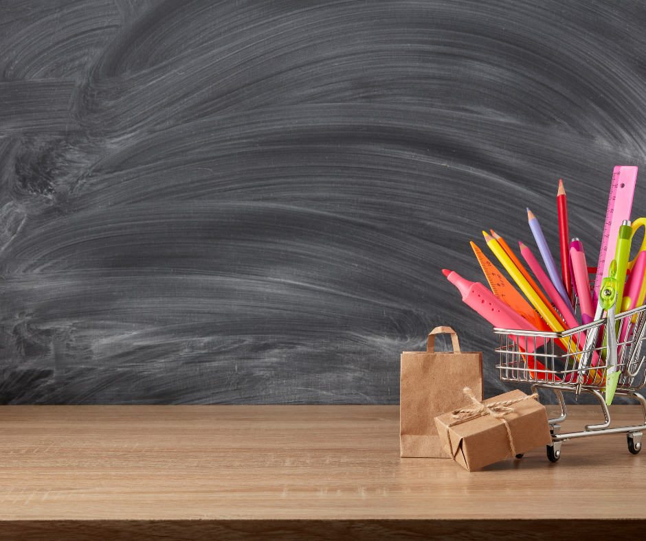 Na całości grafiki znajduje się tablica szkolna, na jej tle umieszczony jest wózek sklepowy wypełniony przedmiotami szkolnymi
