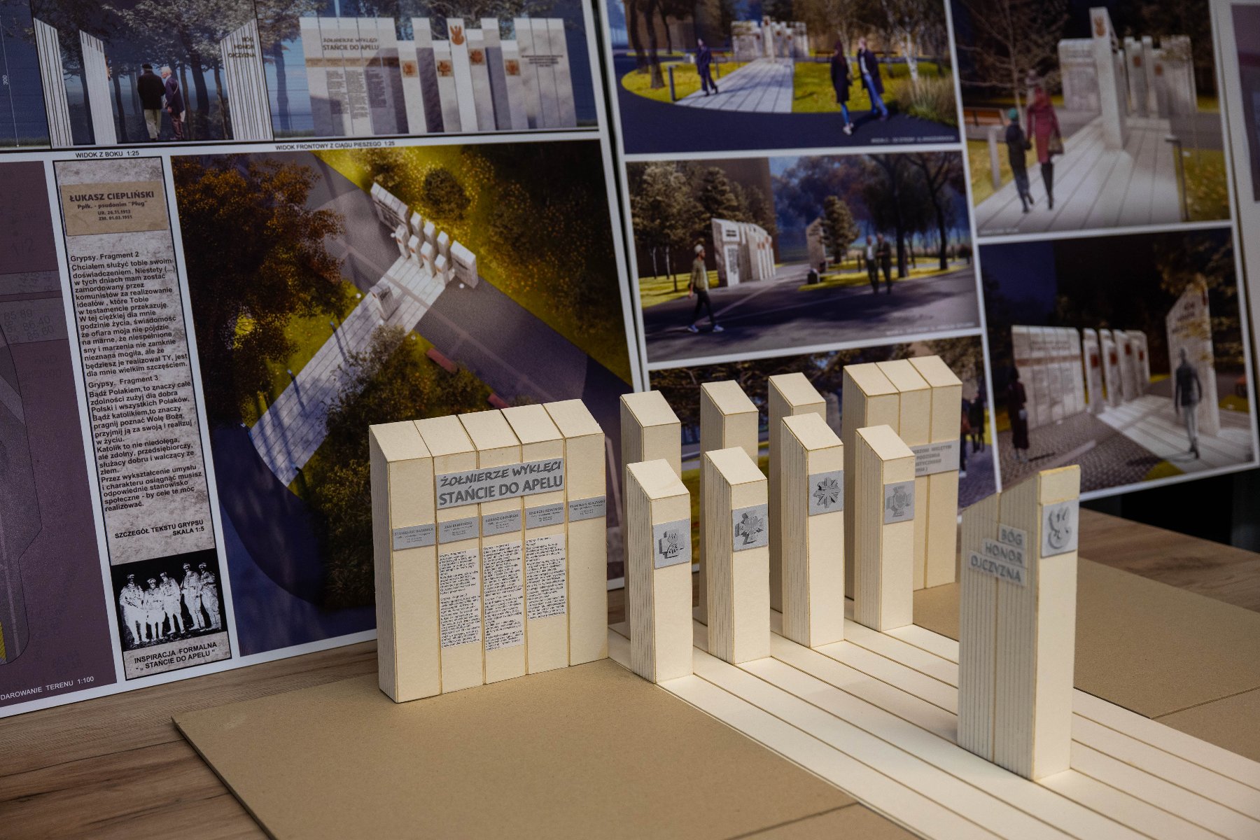 Na drewnianym stole leży plansza z wizualizacjami i tekstem, a przed nią stoi przestrzenna makieta pomnika. Makieta składa się z kilku beżowych prostopadłościanów symbolizujących kamienne tablice.
