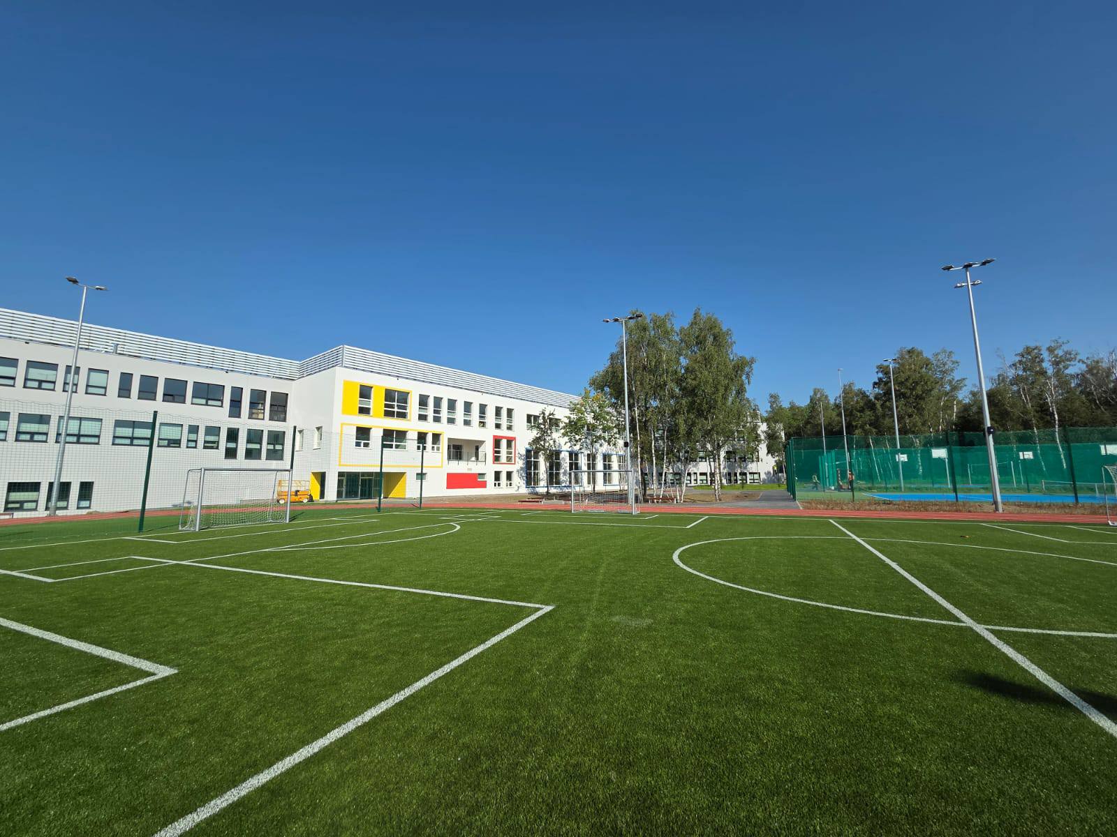 Na grafice widoczne jest trawiaste boisko szkolne do piłki nożnej, a w tyle budynek nowej szkoły.