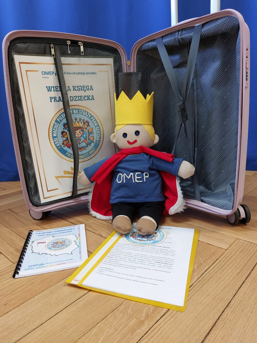 Na grafice znajduje się maskotka przypominająca Króla Maciusia I, siedzi na tle otwartej walizki