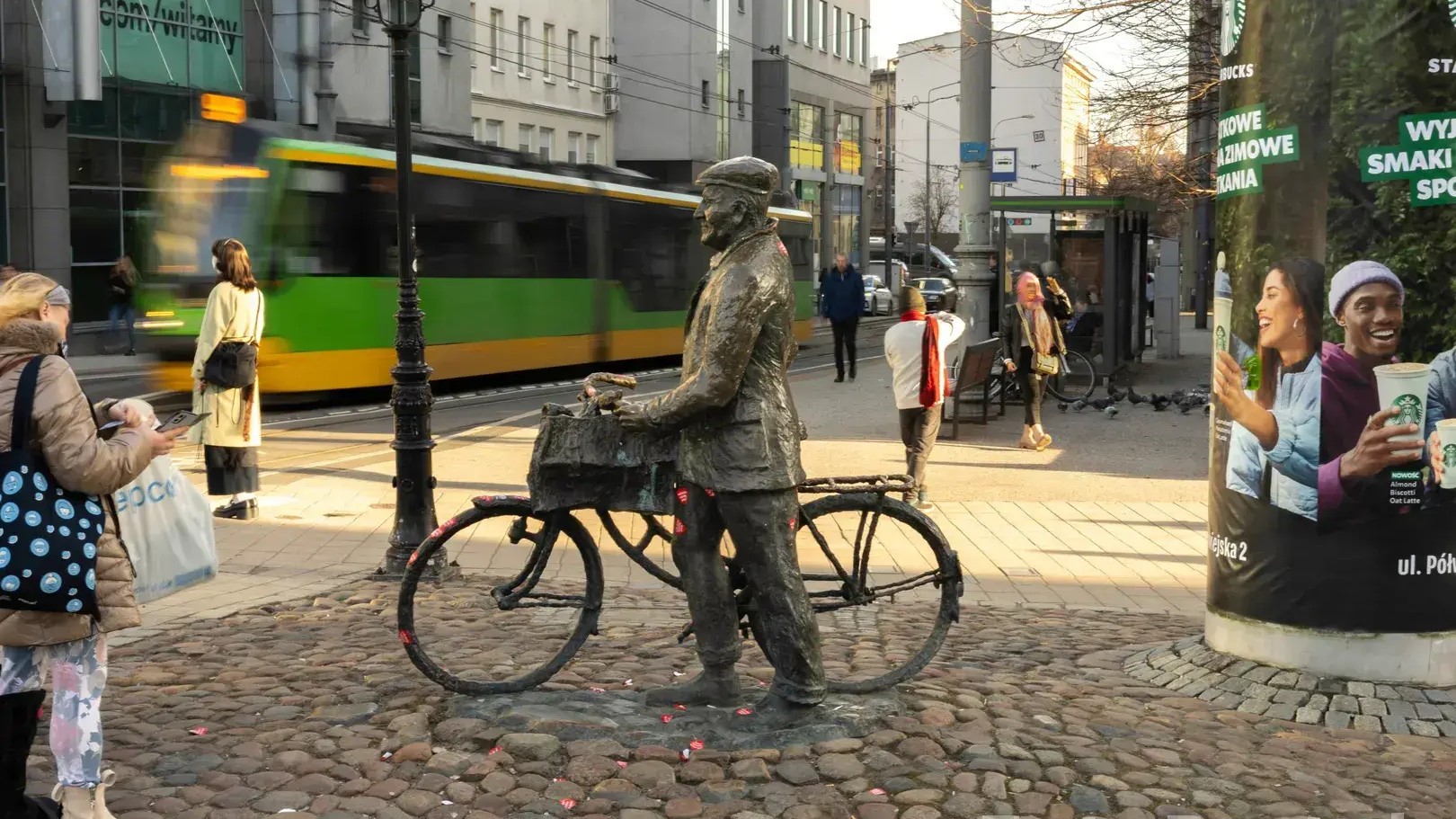 Na miejskim placu stoi pomnik Starego Marycha przedstawiający osobę prowadzącą rower z koszem z przodu. W tle widać przejeżdżający zielono-żółty tramwaj, przystanek komunikacji miejskiej oraz pieszych poruszających się po chodniku między zabudową śródmieścia.