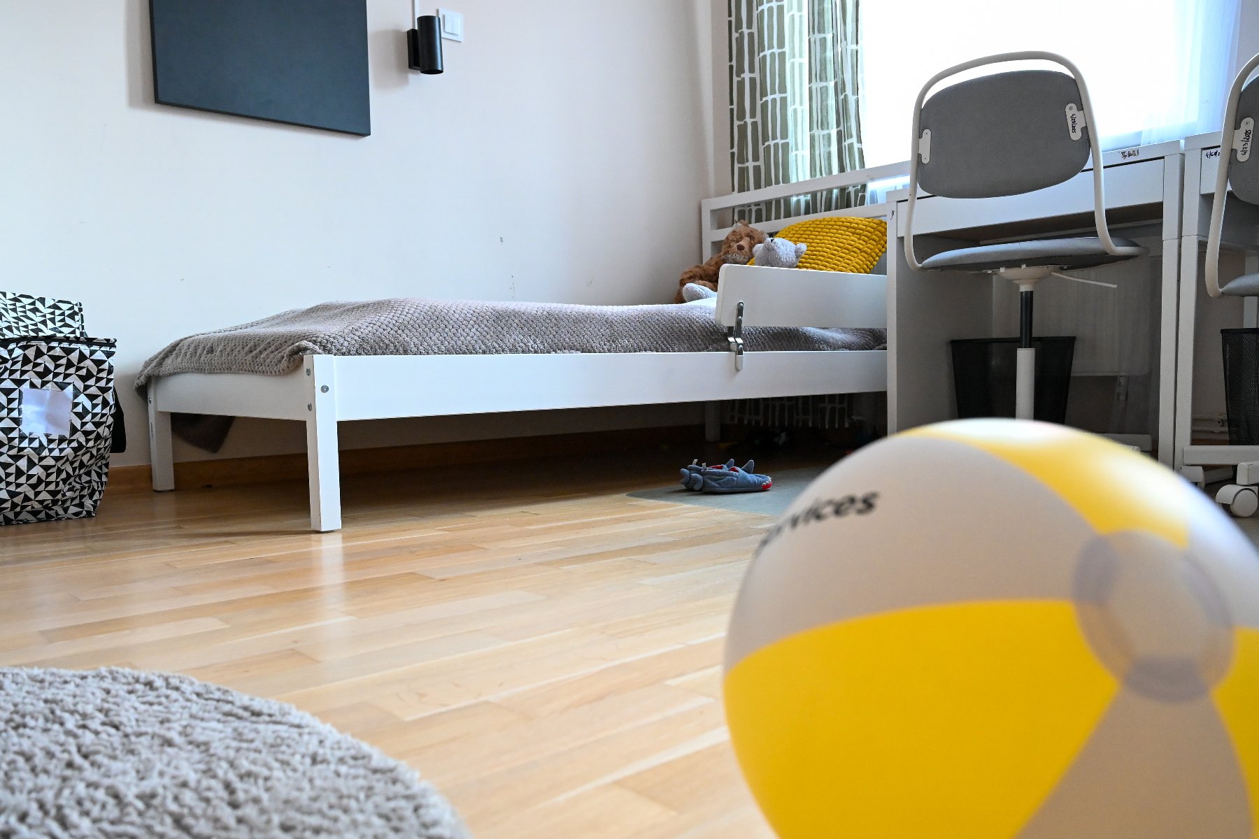 Na obrazku widoczny jest pokój dziecięcy w kolorach żółto szarych. W tle łózko, pod oknem biurko i krzesełko. Na pierwszym planie dmuchana piłka plażowa.
