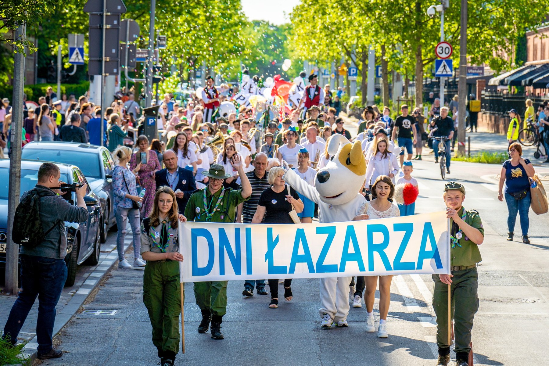 Na pierwszym planie idzie grupa harcerzy i trzyma baner "Dni Łazarza". Za nimi idzie reszta korowodu, która składa się z mieszkańców Poznania. Po drodze stoją inni ludzie, którzy przyglądają się wydarzeniu.