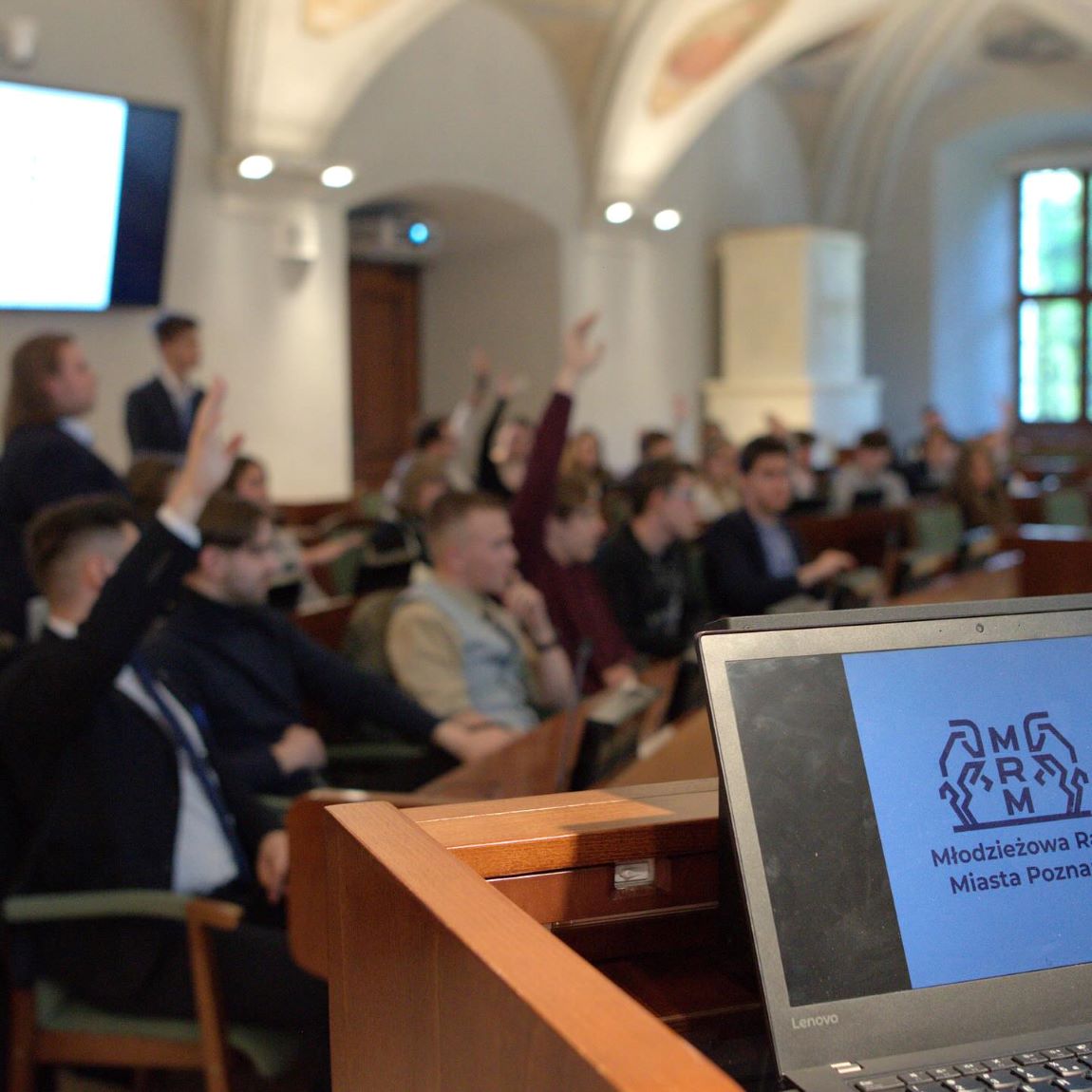 na pierwszym planie otwarty laptop postawiony na mównicy, na ekranie wyświetlone logo i napis Młodzieżowa Rada Miasta Poznania, w tle - zdjęcie zamglone - osoby siedzą przy stanowiska, niektóre z nich mają rękę uniesioną w górę, prawdopodobnie głosują