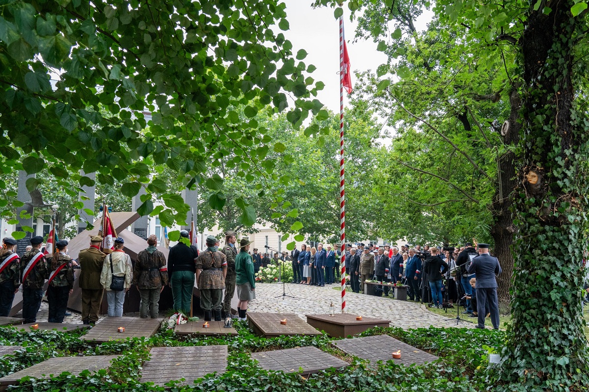 na placu przed pomnikiem odbywają sie uroczości z udziałem harcerzy i oficielu, na placu masz z biało czerwoną flagą, plac otaczają drzewa
