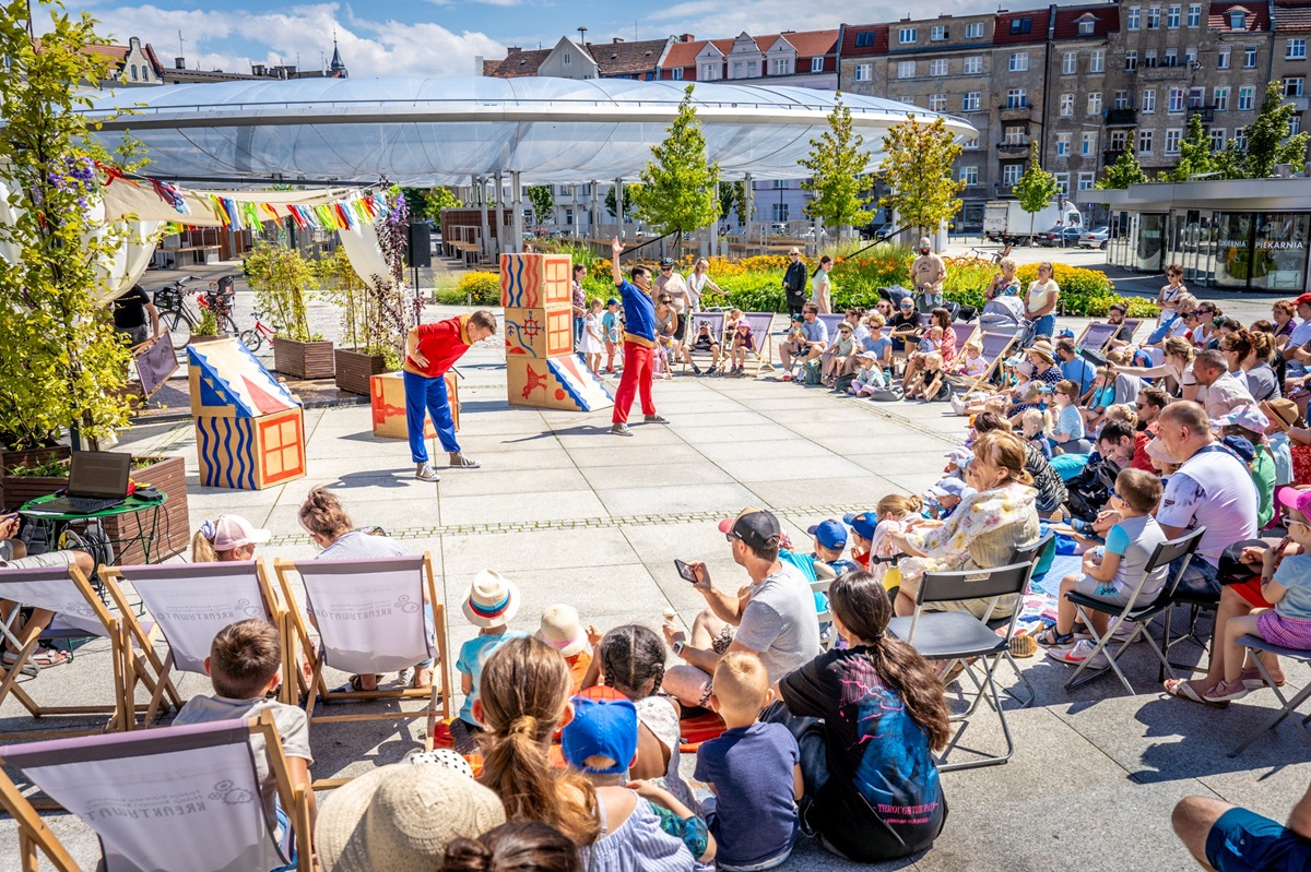 Na płycie Rynku Łazarskiego dwóch mężczyzn - aktorów występuje przed zgromadzoną publicznością. W tle widać kopułę zadaszenia rynku i kamienice.