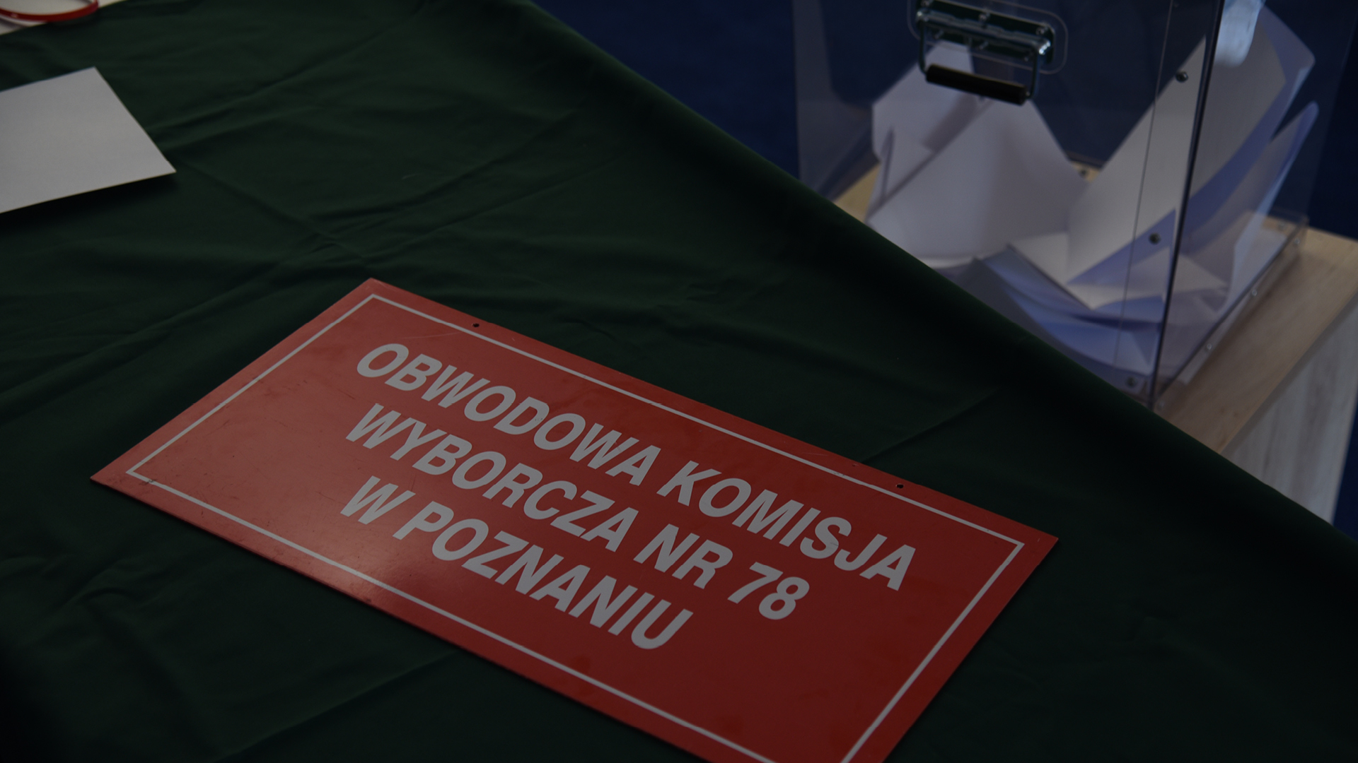 na stole nakrytym zielonym suknem leży czerwona prostokątna tablica informacyjna z białym napisem obwodowa komisja wyborcza nr 78 w Poznaniu. W tle widać wypełnioną do połowy głosami wyborczymi przeźroczystą urnę wyborczą.