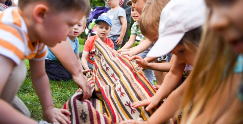 Na świeżym powietrzu grupka dzieci trzyma kolorowy, wzorzysty koc lub dywan. Widoczny pasy kolorystyczne w tonacjach czerwonej, żółtej i brązowej oraz geometryczne i zwierzęce motywy. Dzieci wydają się być zaangażowane w aktywność na świeżym powietrzu, w przestrzeni przypominającej park lub ogród.