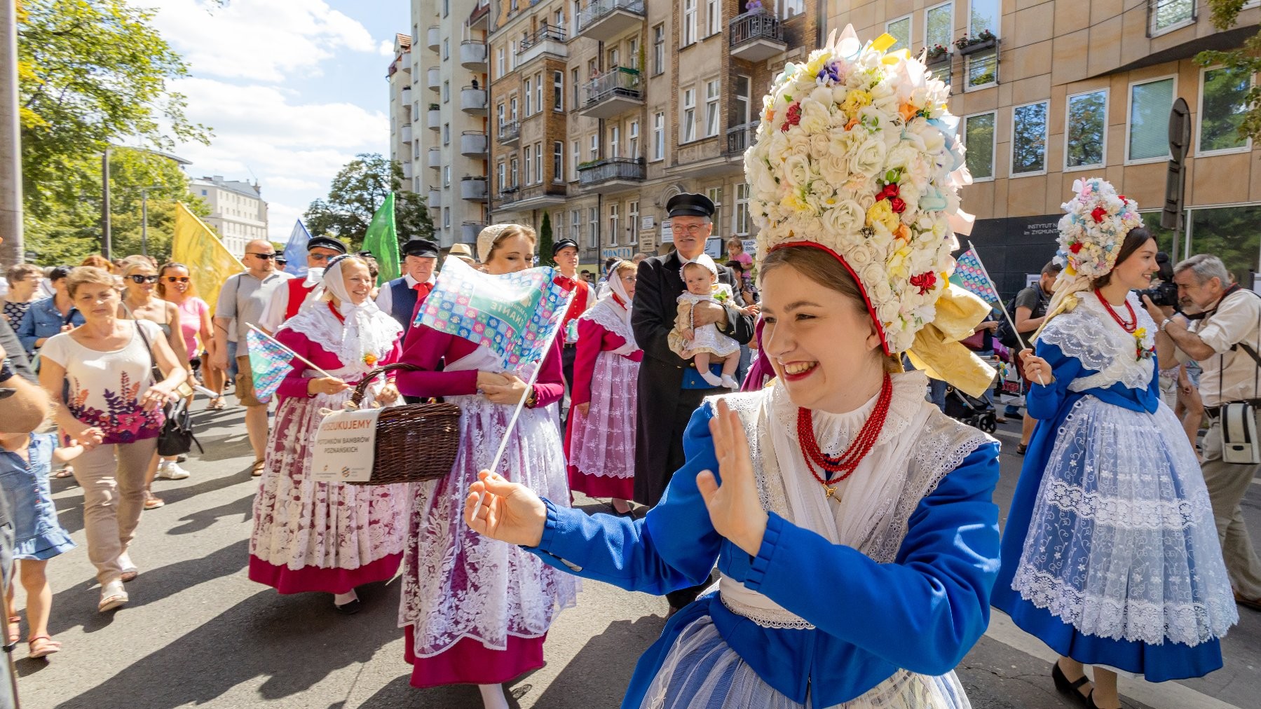 Na ulicy odbywa się barwny pochód. Uczestnicy prezentują tradycyjne stroje bamberskie charakterystyczne dla potomków osadników bamberskich związanych z Poznaniem. Na pierwszym planie widoczna jest osoba w niebiesko-białym stroju z czerwonymi koralami i wysokim kwiatowym czepcem. Wokół niej idą inni uczestnicy w bogato zdobionych strojach bamberskich, w otoczeniu licznych widzów. Widać też kamienice i drzewa skąpane w słonecznym świetle.