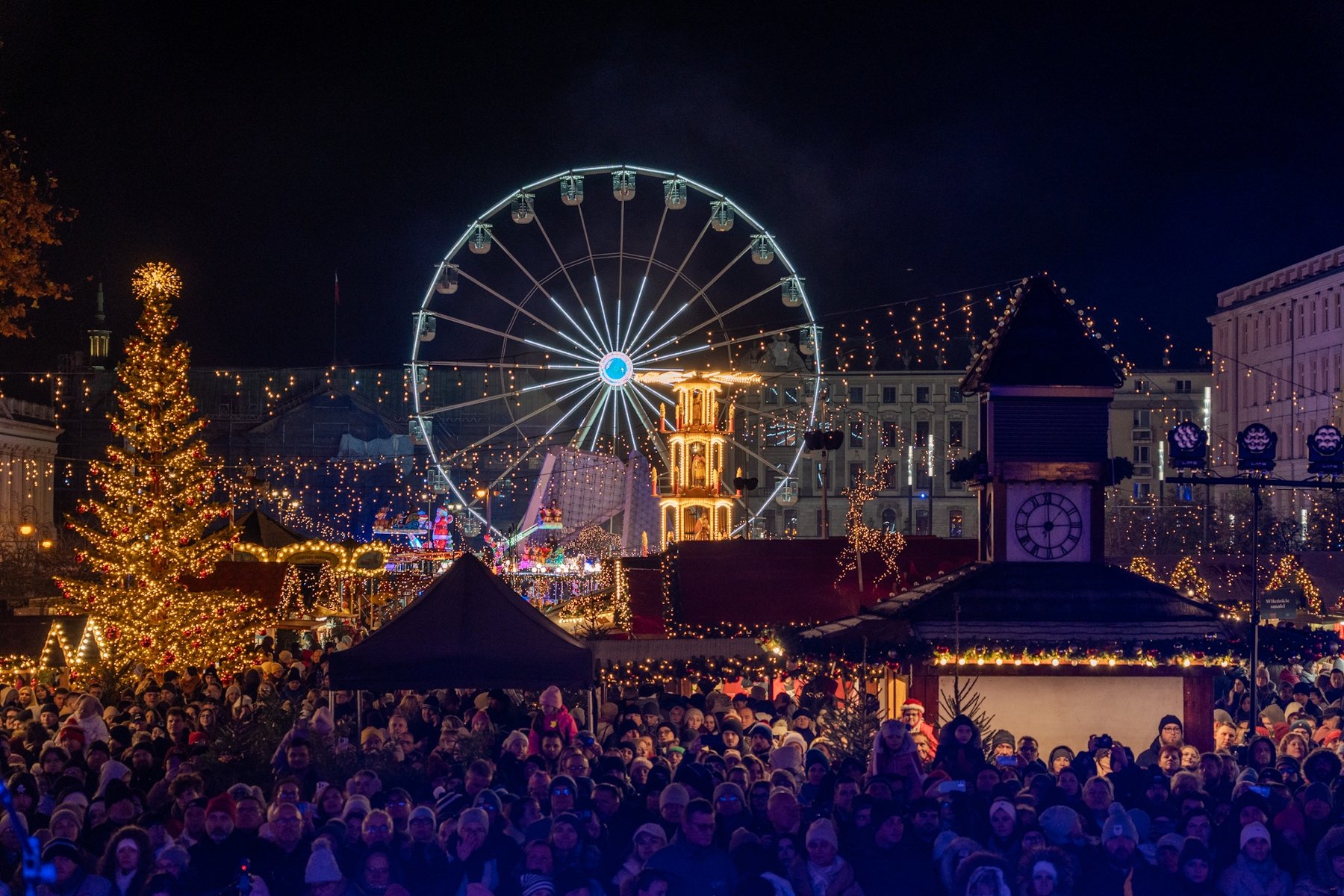 Na zdjęciu alejka na jarmarku, widać młyńskie koło i stoiska, między nimi spacerują ludzie