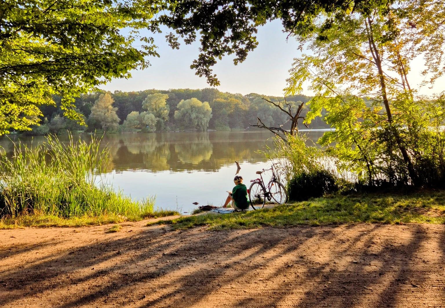 Na zdjęciu brzeg jeziora, nad nim siedzi kobieta, obok stoi rower