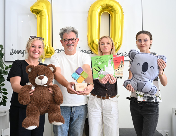 Na zdjęciu cztery osoby, trzy kobiety i mężczyzna, uśmiechają się do obiektywu, w rękach trzymają zabawki, książki, statuetkę. W tle balony w kształcie liczby 10