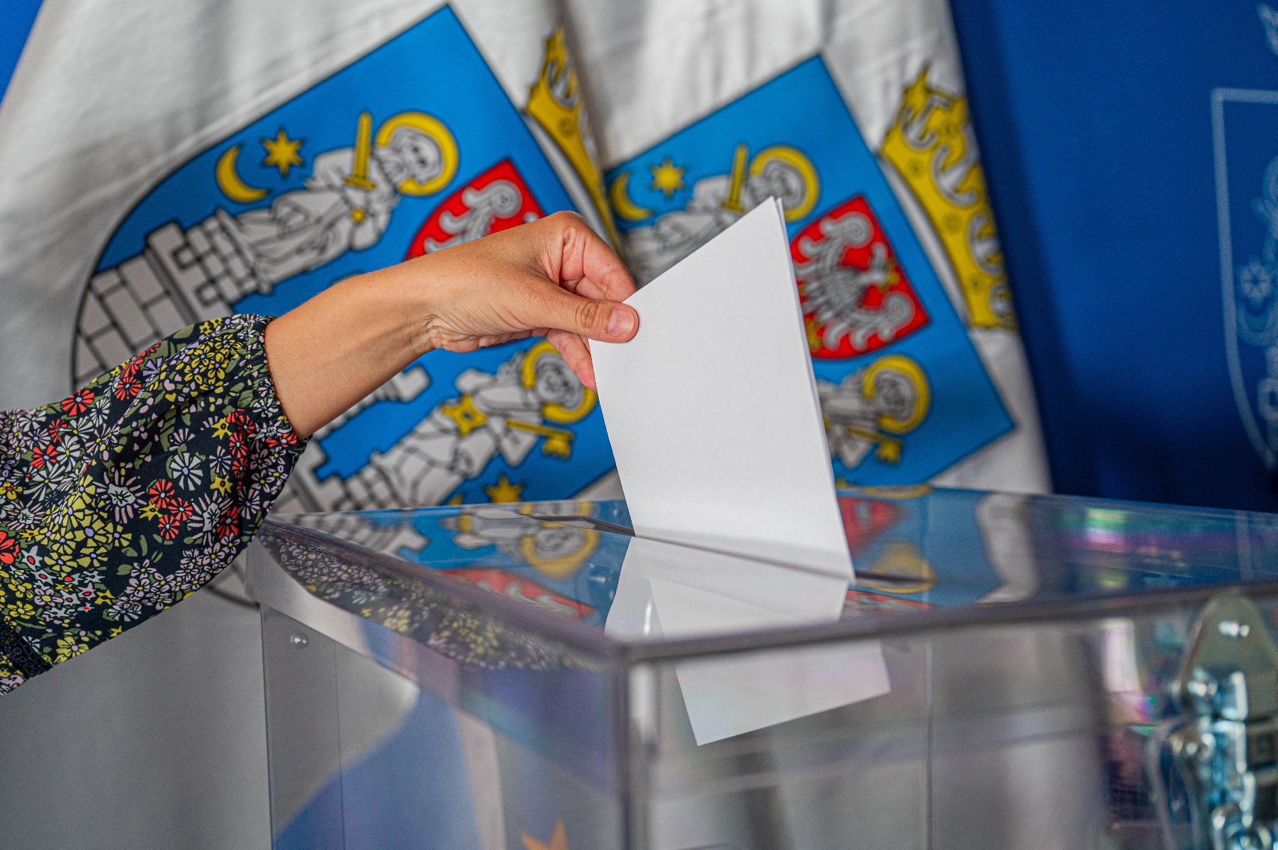 Na zdjęciu dłoń wrzucająca głos do urny, w tle dwie flagi miasta