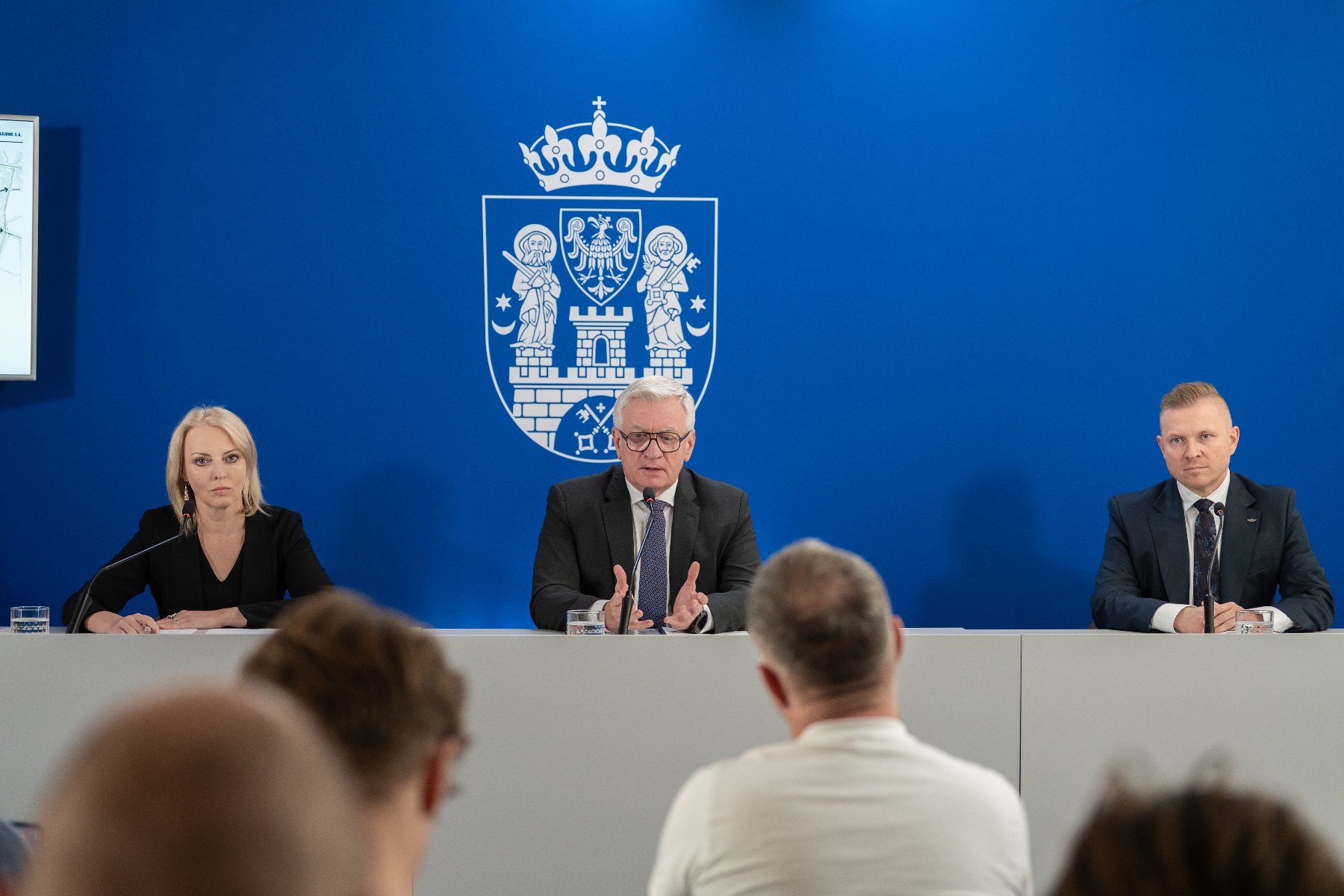 Na zdjęciu dwaj mężczyźni i kobieta, za nimi ścianka z herbem miasta