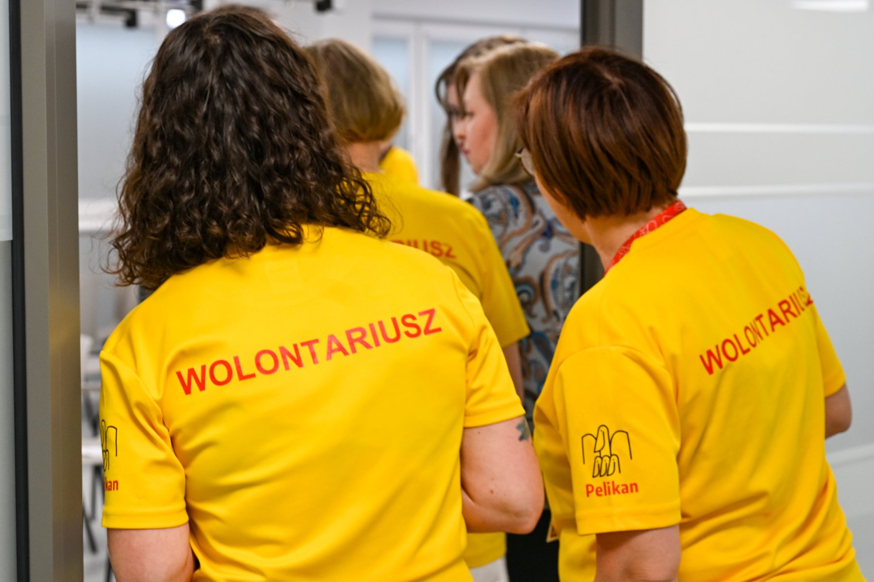 Na zdjeciu dwie kobiety stojące tyłem, na żółtych koszulkach mają napis: wolontariusz