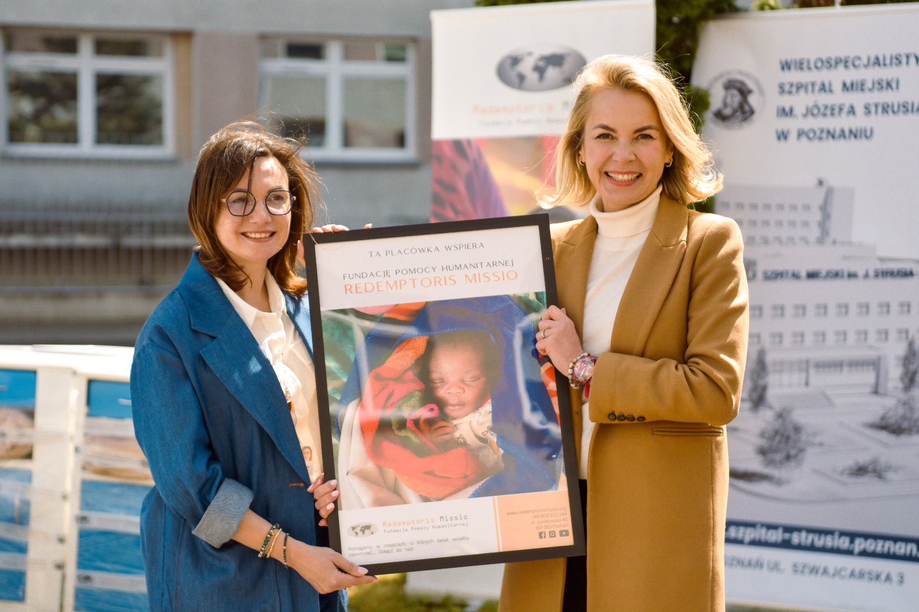 Na zdjęciu dwie kobiety trzymające plakat, na nim małe dziecko