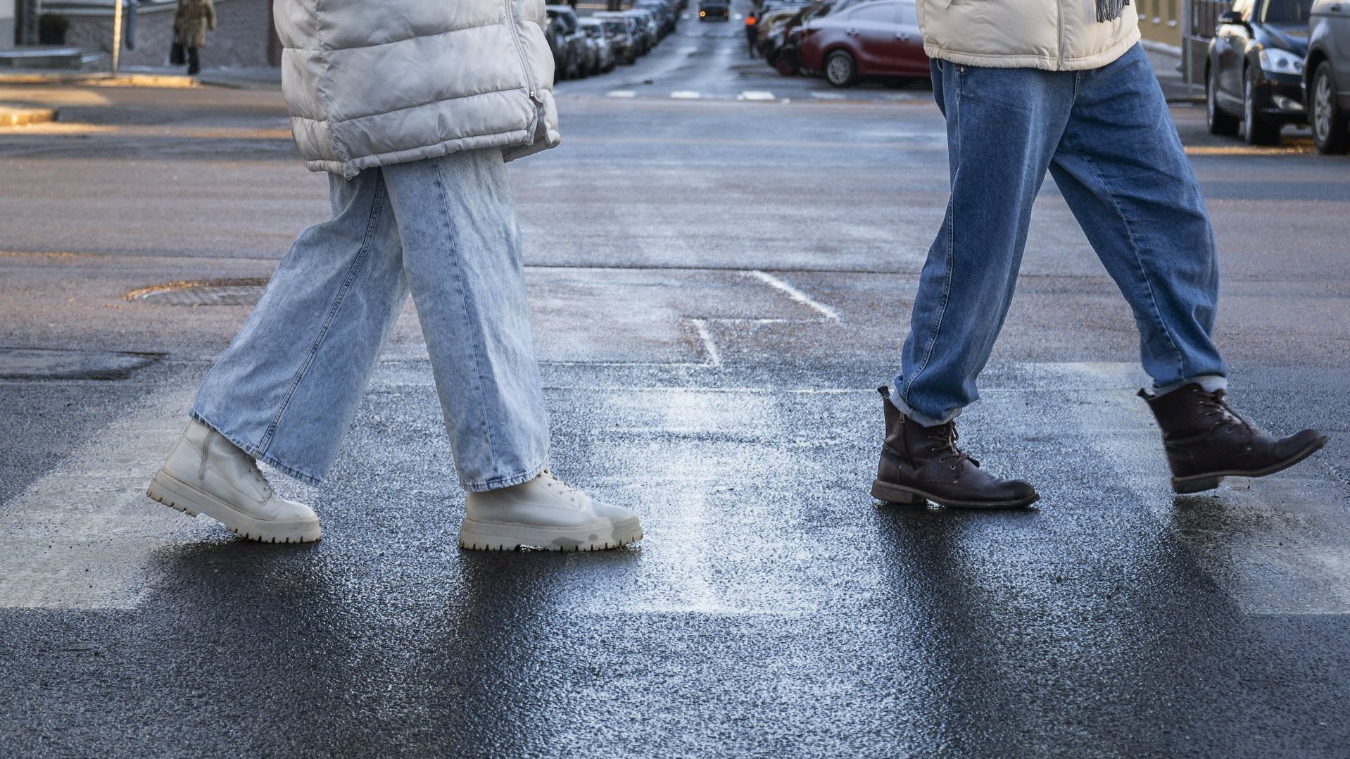 Na zdjęciu dwie osoby przechodzące przez przejście na pasach na wilgotnej jezdni, widać tylko ich nogi