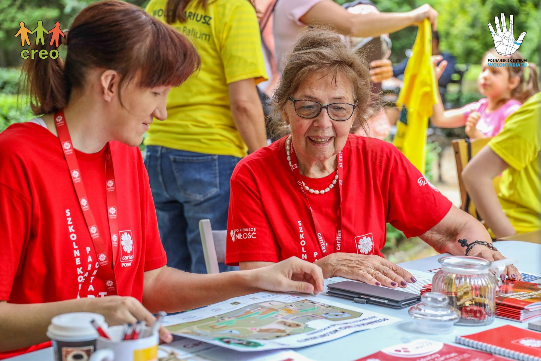 Na zdjęciu dwie wolontariuszki w czerwonych koszlkach siedzące przy stoliku, w tle dzieci