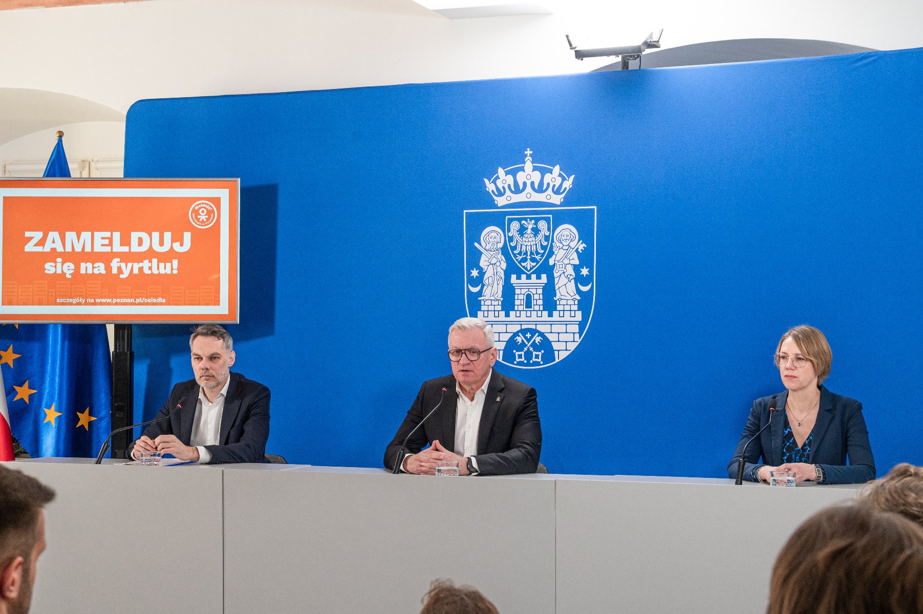 Na zdjęciu dwóch mężczyzn i kobieta za stołem konferencyjnym, w tle niebieska ścianka z herbem miasta
