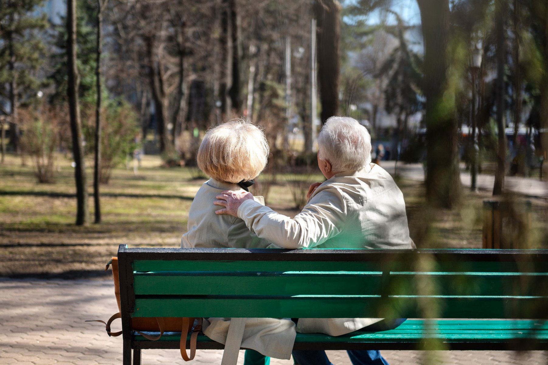 Na zdjęciu dwoje seniorów siedzących na ławce w parku, tyłem do obiektywu