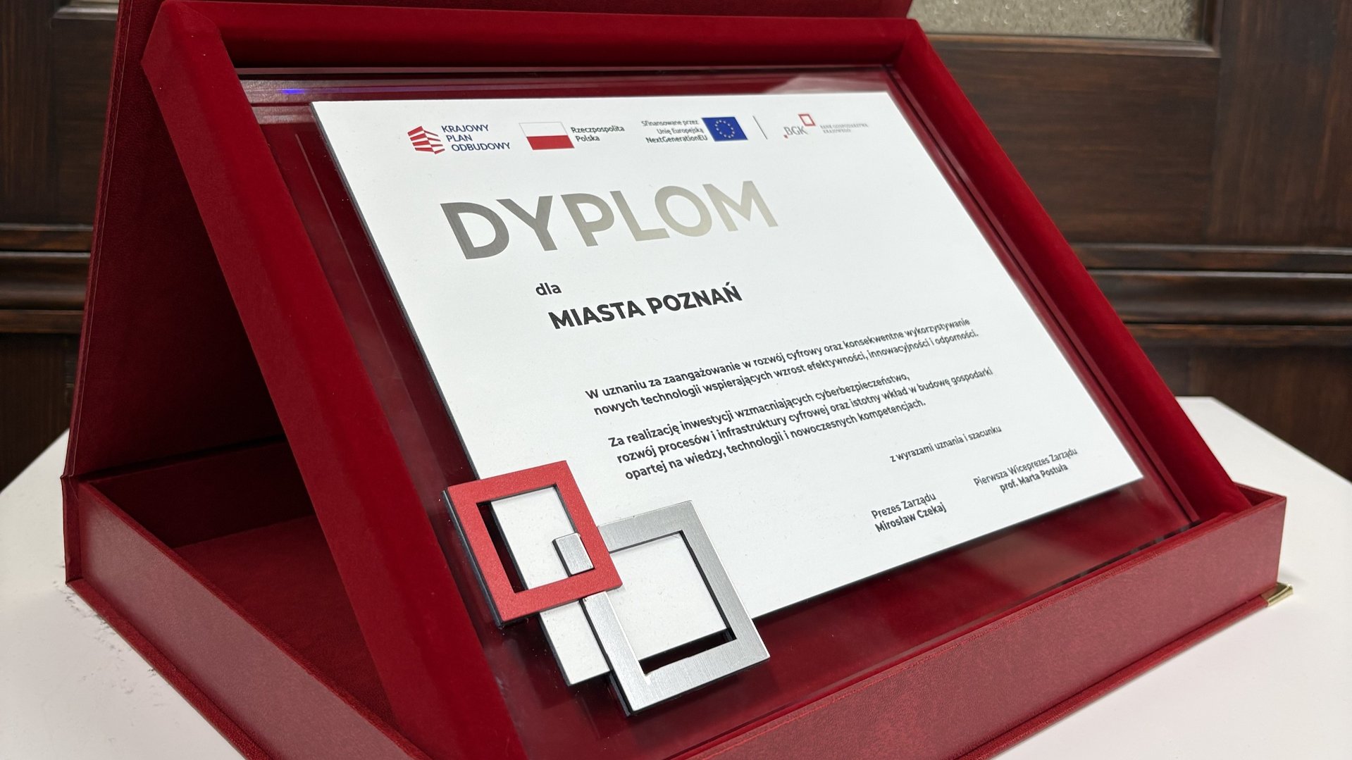 Na zdjęciu dyplom dla Miasta Poznania przyznany przez Bank Gospodarstwa Krajowego w uznaniu za zaangażowanie w rozwój cyfrowy oraz konsekwentne wykorzystywanie nowych technologii wspierających wzrost efektywności, innowacyjności i odporności. Za realizację inwestycji wzmacniających cyberbezpieczeństwo, rozwój procesów i infrastruktury cyfrowej oraz istotny wkład w budowę gospodarki opartej na wiedzy, technologii i cyfrowych kompetencjach. Dyplom stoi w etui na stole. - grafika artykułu