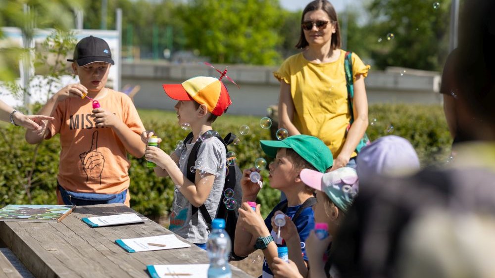 na zdjęciu grupa dzieci siedząca przy stole, puszczają bańki mydlane. Za nimi stoi kobieta, obserwuje bawiące się dzieci.