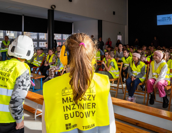 Na zdjeciu grupa dzieci w kamizelkach odblaskowych, przyglądają się czemuś