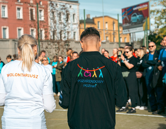 Na zdjęciu grupa ludzi, dwie osoby stoją tyłem, na koszulkach mają napisy: wolontariusz, pozarządowy Poznań