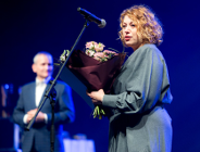 Na zdjęciu kobieta trzymająca kwiaty mówi coś do mikrofonu